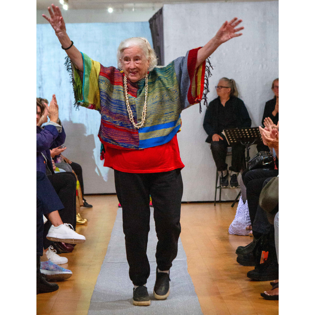 a woman with light skin and white hair wearing a handwoven poncho walks down the runway, arms raised.