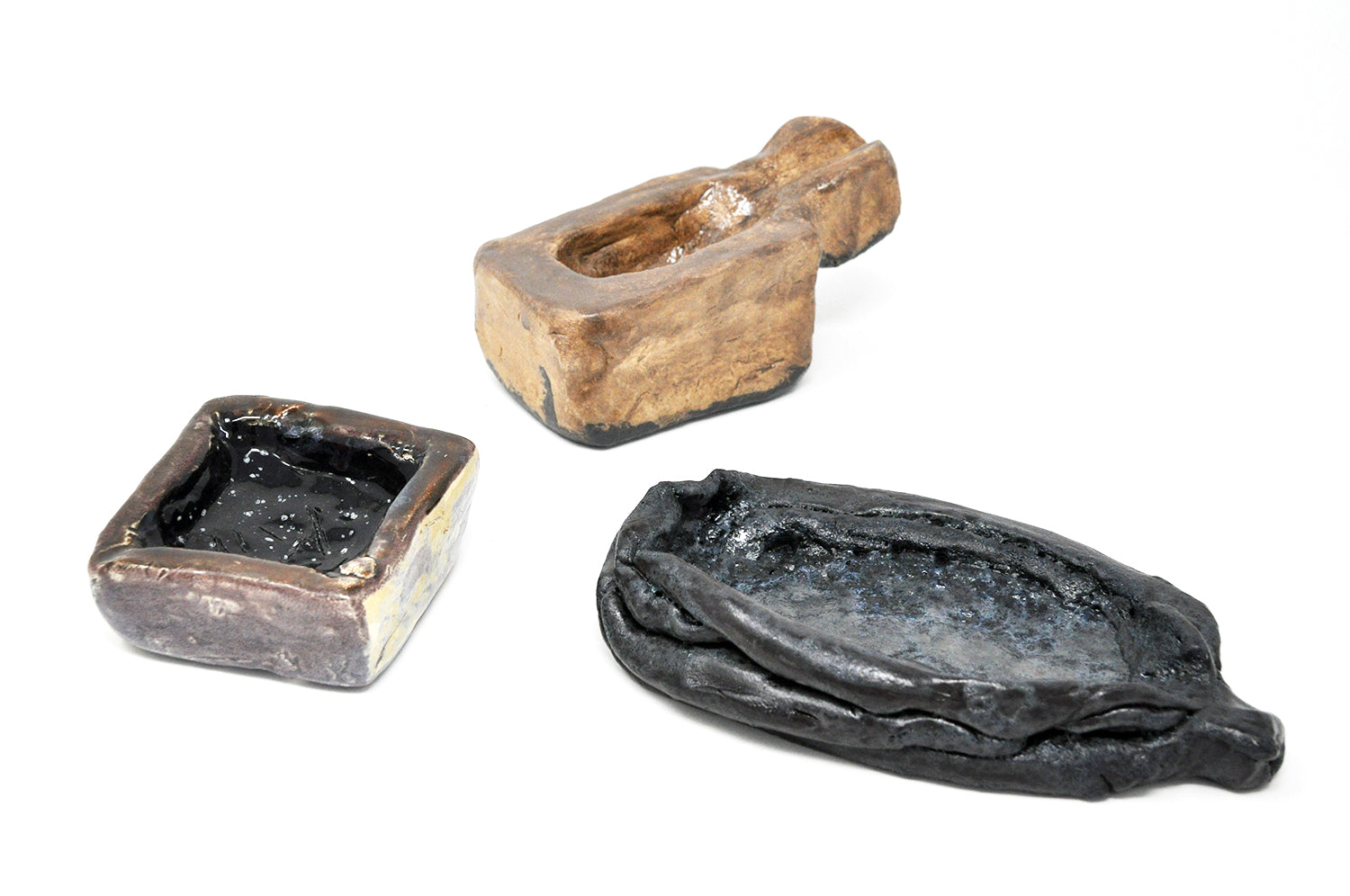 three clay ceramic sculptures laid in a circular formation viewed from above. Each piece varies in rectangular shape and is glazed a brown or black color. All pieces have a shallow opening in the center. 