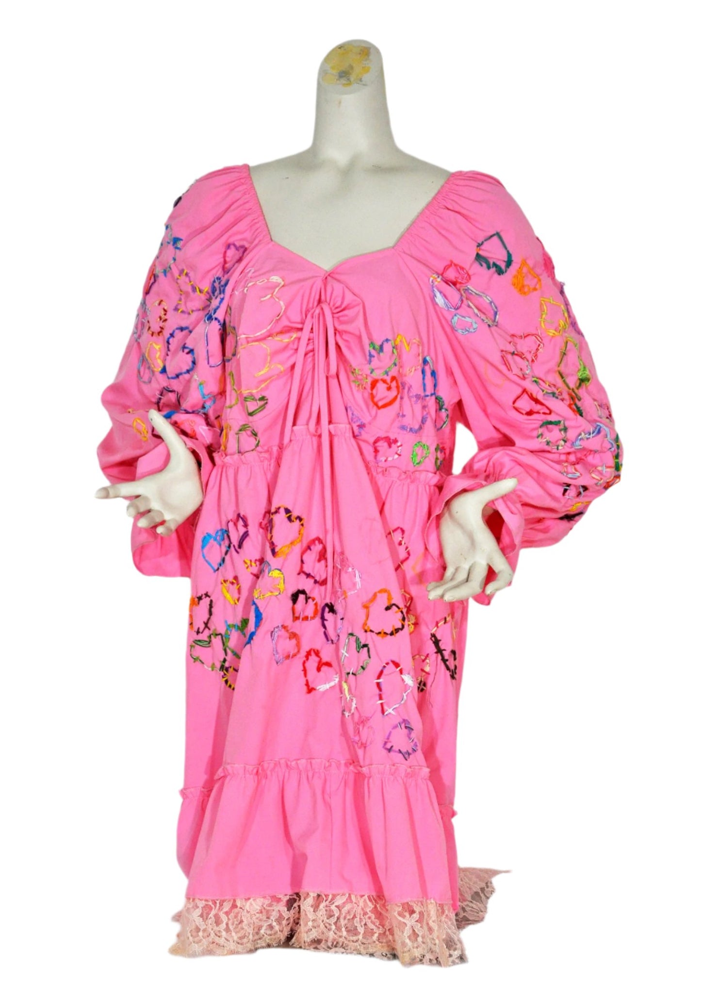 a short pink dress with long sleeves modeled on a grey manikin torso, photographed against a stark white backdrop. The front of the dress has been embroidered with rows of hearts in every color of the rainbow. There is pale pink lace trim stitched along the bottom hem.