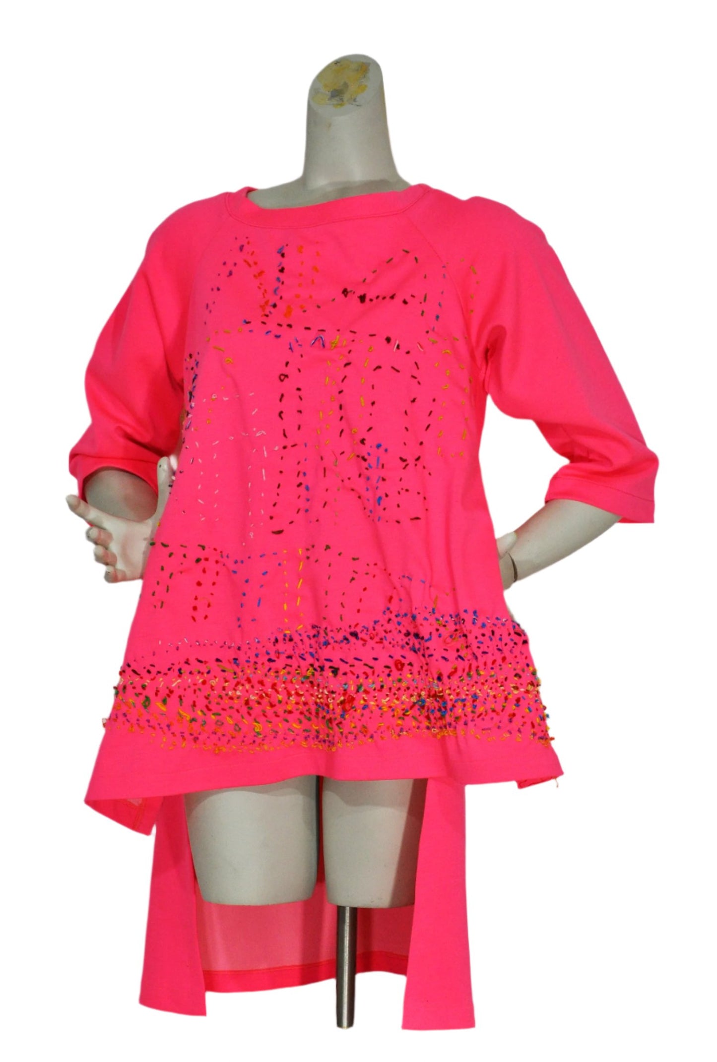 a bright pink tunic modeled on a grey manikin torso. The tunic has been embroidered all over with delicate dashed stitches using thread in every color of the rainbow. Many stitches cluster to form a wide rainbow band around the bottom hem of the garment. In the same running stitch style, the artist has also embroidered the letters "NIAD" across the chest.  