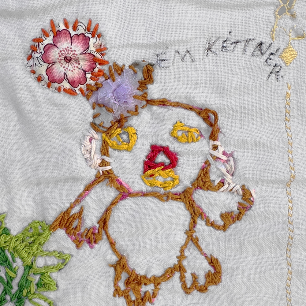 a detail photograph of an embroidered textile. On a pale grey woven cloth, a little figure is embroidered, outlined with brown thread. The figure has stitched yellow eyes, a red nose, and yellow mouth. The figure is wearing a stack of purple and pink appliquéd flowers as a hat.  The name "Em Kettner" is written in black fabric marker next to the figure. 