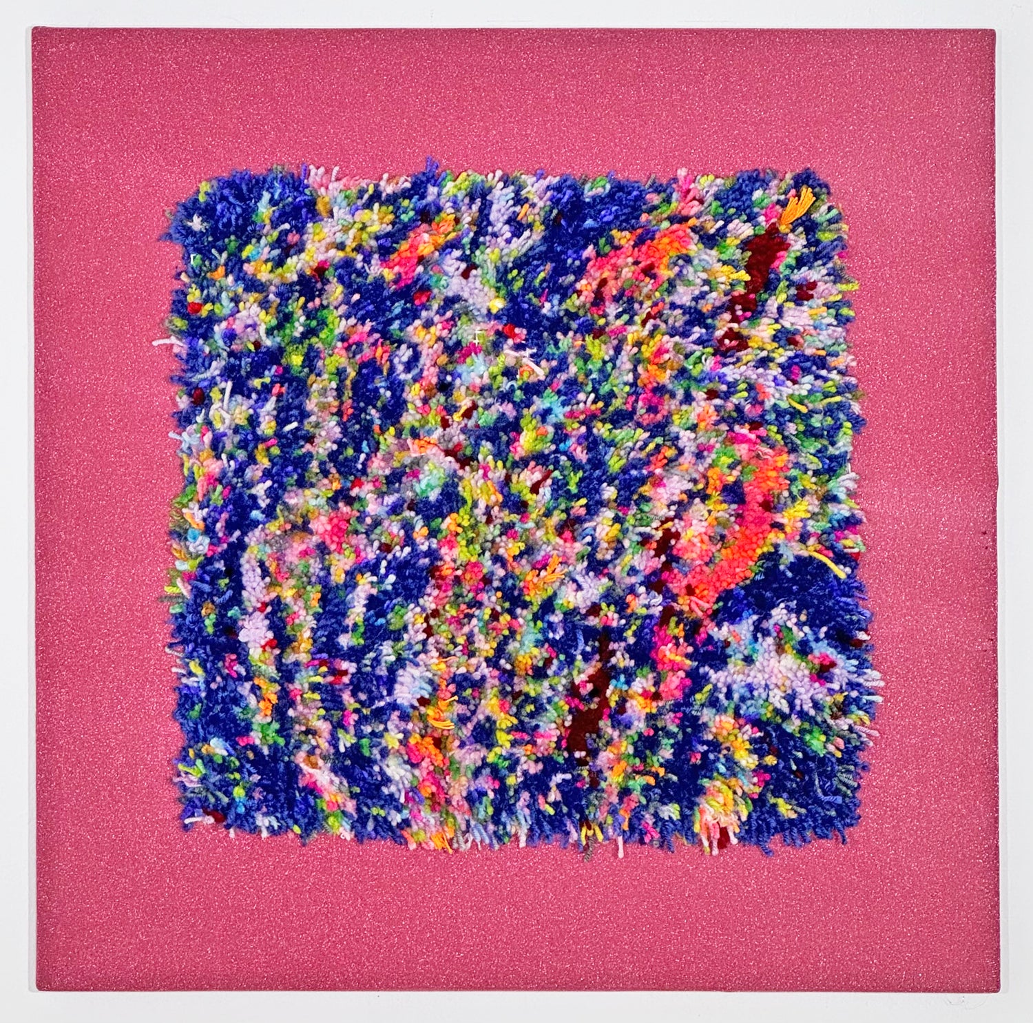 A tufted rug attached to the center of a bright pink piece of fabric. The fabric has been stretched over stretcher bars so that the piece can hang like a painting on the wall. The tufted rug is a square shape made of short pieces of yarn. The surface features an abstract striped field of blue, white, and pink, with small flecks of every color of the rainbow peppered around the surface. 