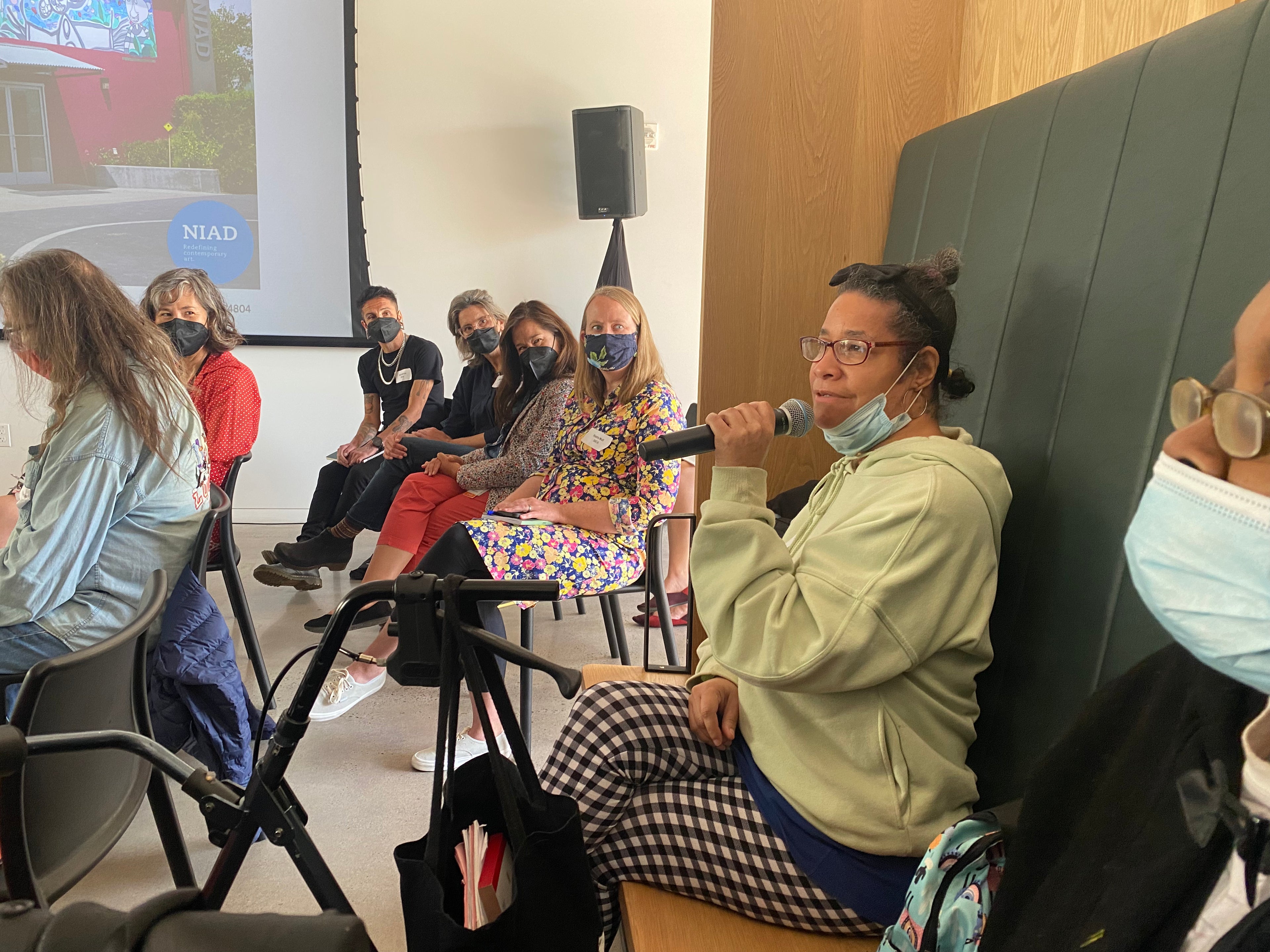 A photograph of a crowd of people sitting indoors. The focus of the image is a woman sitting in the foreground on the right: the artist Deatra Colbert. Deatra has brown skin and short black hair pulled into a bun. She wears glasses, a green sweatshirt, and black and white checkered pants. She holds a microphone. The people in the surrounding crowd are all wearing masks and looking at Deatra while she speaks. There is a projector screen on the wall in the background showing an image of the NIAD building.  