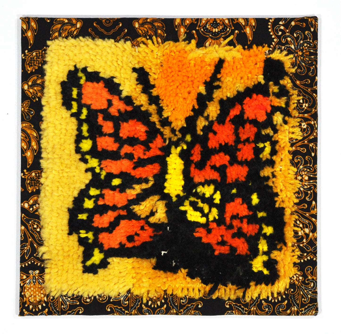 A fiber work stretched over square stretcher bars. The work is made of tufted yarn attached to an ornately patterned piece of black and orange cloth. The tufted section depicts an orange and yellow butterfly on a bright yellow surface. 