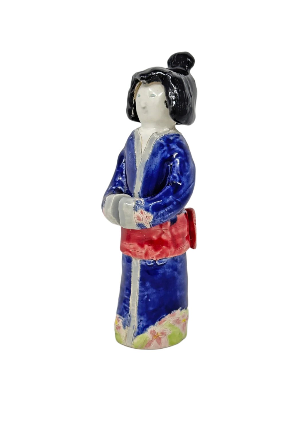 a small ceramic figurine dressed in traditional Japanese clothing and glazed blue, white, and red. The garment is a long blue robe with a floral design at the bottom hem, and a wide red sash tied around the waist. The figure's hands hidden by long sleeves, but appear joined together at the figure's front in a relaxed position. The figure has black hair pulled into a loose bun.