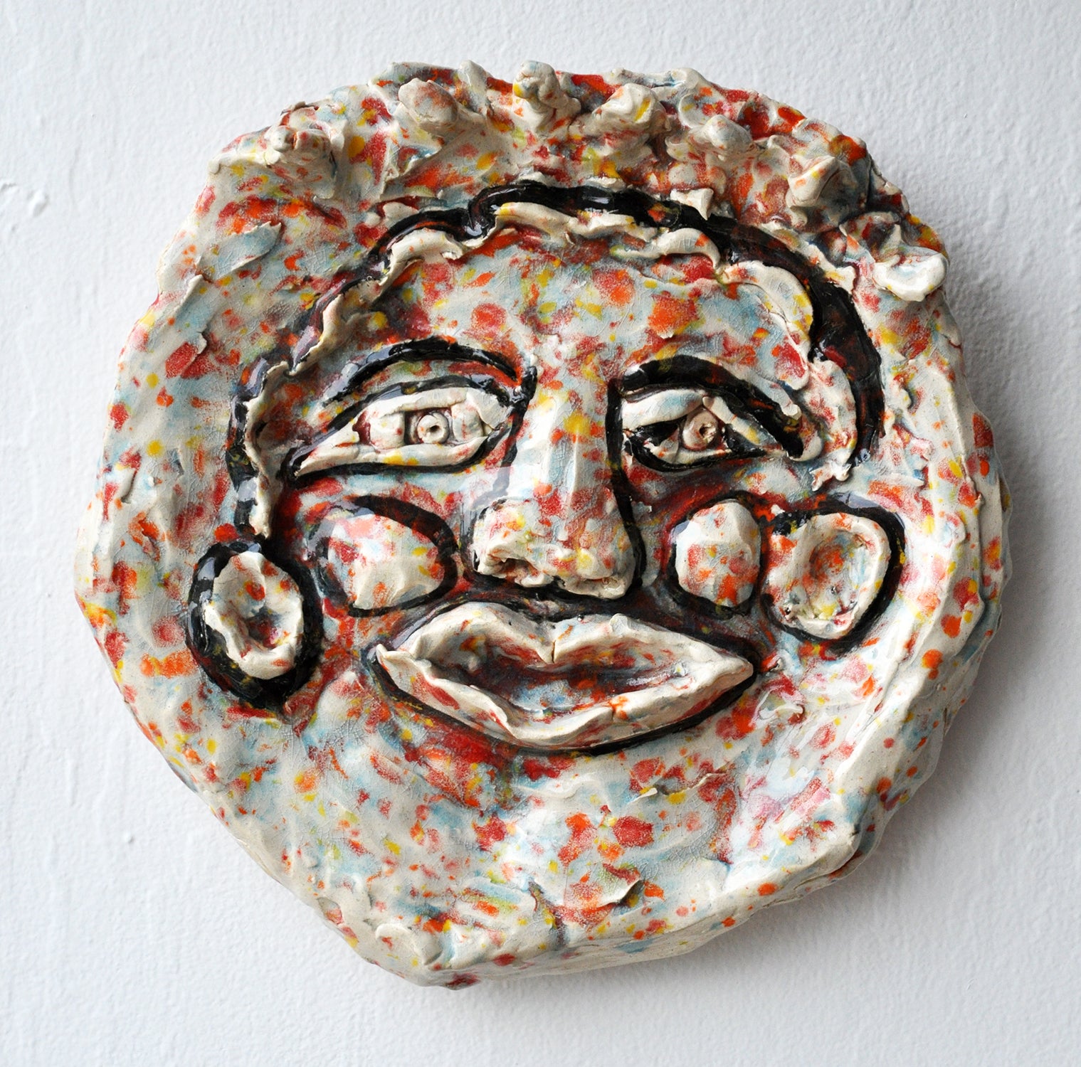 A face with features outlined in black glaze. The background glaze colors look like bright scattered confetti in tones of red, yellow, orange, and blue. 