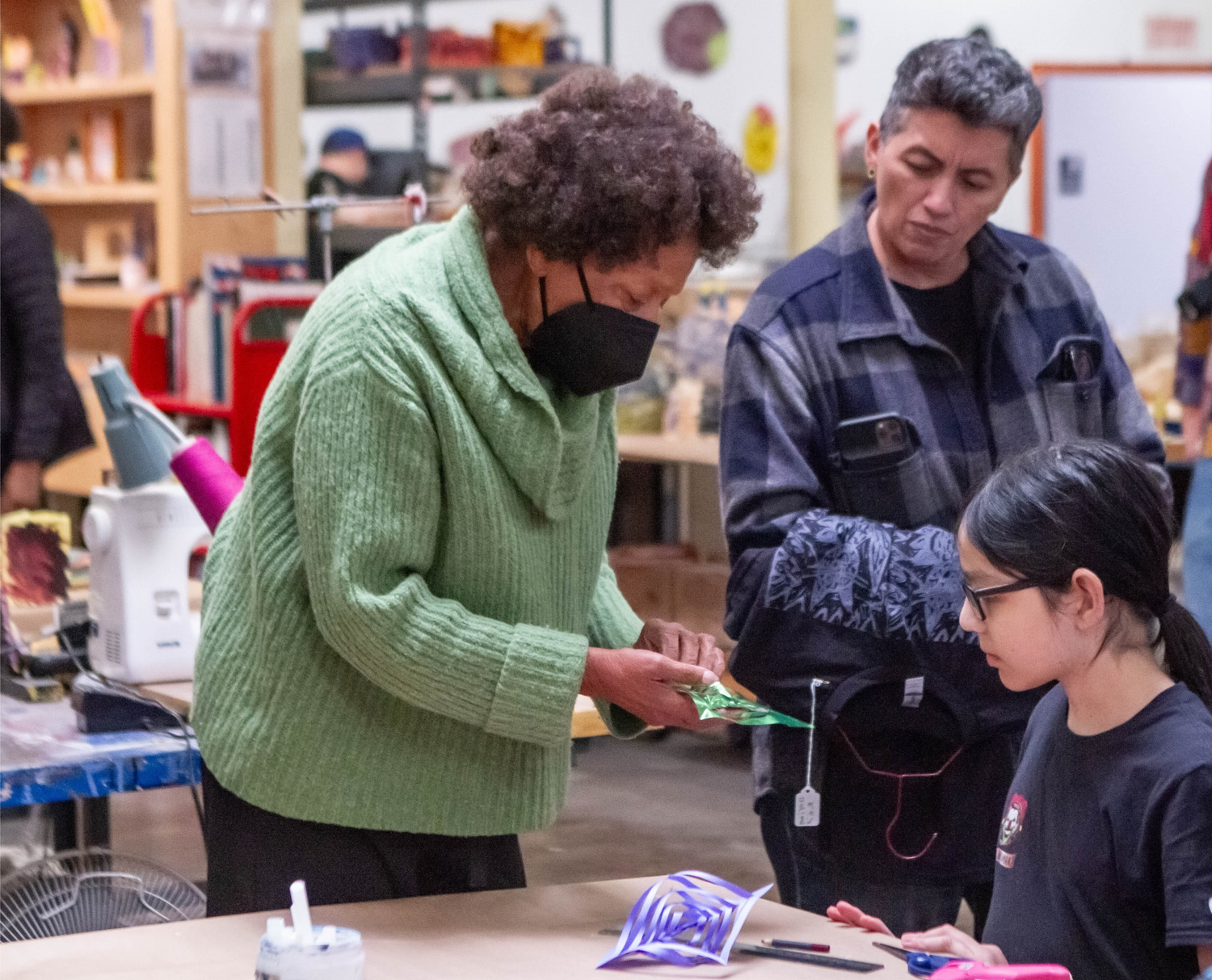 An image of several people in an art studio. In the center of the photo stands a person with short curly brown hair and brown skin. They wear a green sweater and a black mask, and they are showing a green piece of paper to a young person seated at a table. The paper has been cut and folded in an intricate spiral pattern. There is a similarly cut piece of purple paper on the table. Another adult wearing a blue plaid jacket stands in the background watching the demonstration.