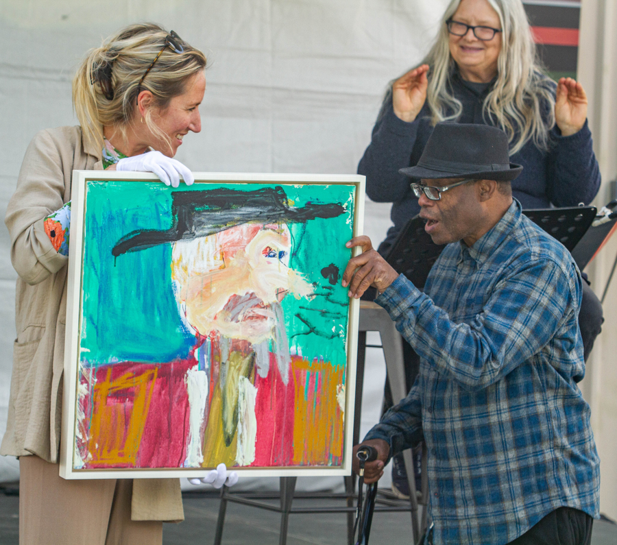 A photograph of three people at an art auction. The figure to the left is a white woman with blond hair, white gloves, and a tan suit. She holds up a portrait painting. The artist, Arstanda Billy White, stands to the right, and gestures at the painting. Arstanda is a black man with glasses, a black hat, and a blue plaid shirt. He carries a cane and has opened his mouth to speak. Seated on the stage behind him is a sign language interpreter: a person with long white hair, light skin, and a blue sweater. 