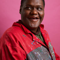 A close-up portrait photograph of a seated person. The person has brown skin, short black hair, and brown eyes. They are looking off into the distance toward the left. They are wearing a red and grey long sleeve shirt splattered with paint. The backdrop is a light purple wall. 