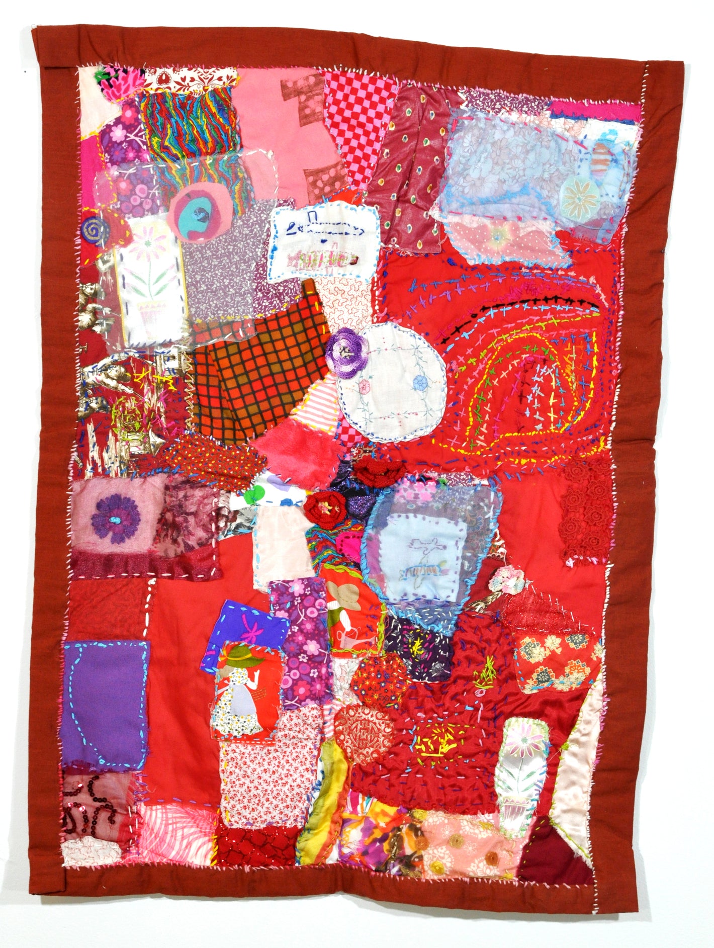 A red quilt photographed hanging on a white wall. The quilt is a tall rectangle with a dark red border. The surface is a layered, hand-stitched collage of red and purple fabric swatches. Many of these swatches have floral patterns. Some sections of the quilt are intricately embroidered with circles and stripes in a rainbow colored running stitch.