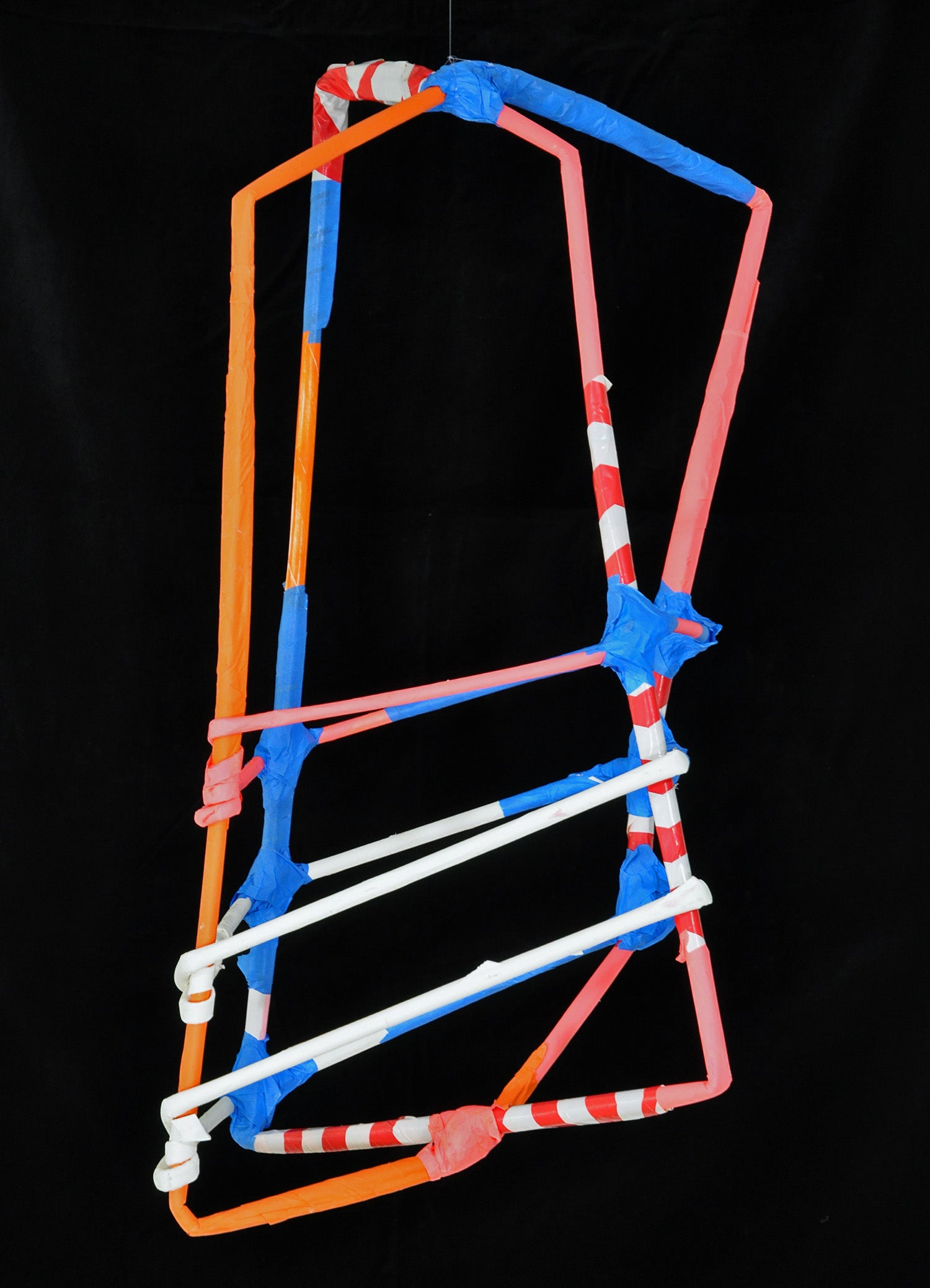 a sculpture made from tightly rolled tubes of paper and tape, photographed against a black backdrop. The form is a cage-like structure, with vertical and horizontal tubes bound by blue or red tape wherever they intersect. The tubes are colored variously orange, red, white, or striped red and white. 