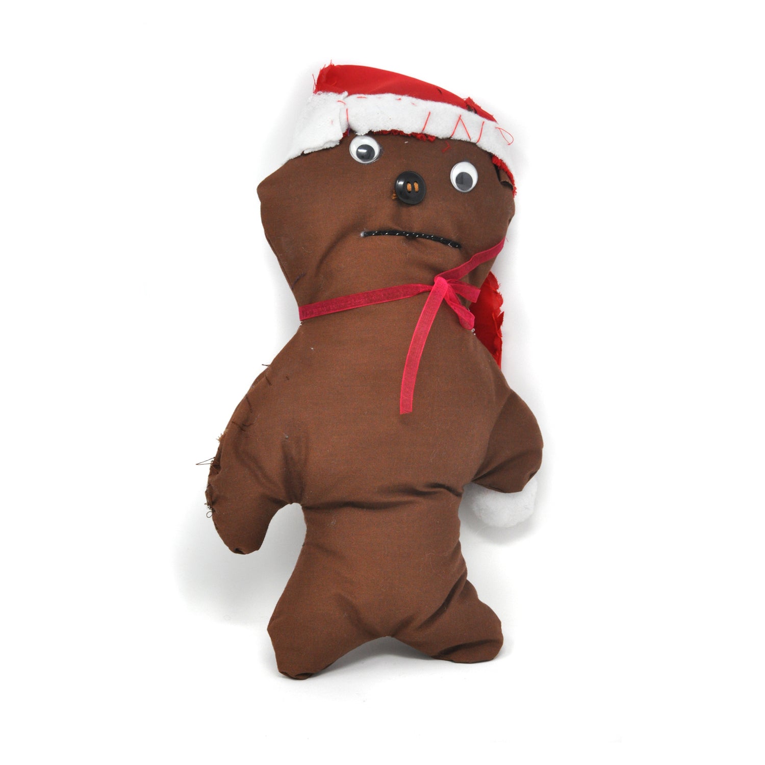 A soft stuffed sculpture photographed against a white backdrop. The sculpture is a small doll figure made out of brown cloth. The head is large and has 2 googly eyes, a black button nose, and a thin closed mouth. The doll is wearing a red pointy hat in the style of Santa Claus, and a red bow around its neck. 